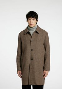 Cappotto a quadri marrone realizzato in tessuto misto lana, con colletto a revers, chiusura con bottoni e tasche laterali. Indossato sopra un dolcevita grigio.