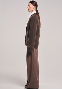 Bruine gebreide cardigan met een losse pasvorm, twee voorkruiszakken en tekstdetail. Combinatie met wijde bruine broeken en zwarte puntige schoenen.