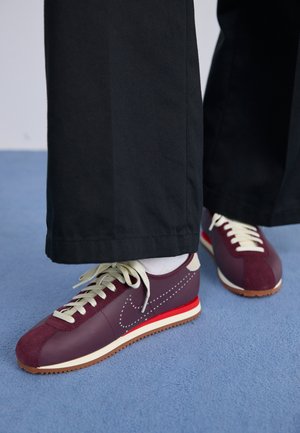 Baskets athlétiques bordeaux et crème avec un mélange de daim et de matière lisse, présentant un accent rouge contrastant, des lacets blancs et une semelle texturée.