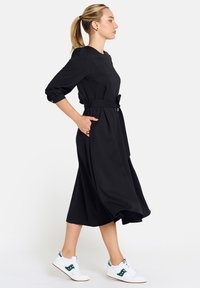 Musta midi-mekko, jossa on 3/4-hihat, vyötärölle vyö ja sivutaskut. Yhdistetty vihreillä yksityiskohdilla varustettuihin valkoisiin tennareihin. Sileä kangas.