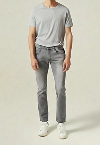 Homme portant un t-shirt gris clair à manches courtes, un jean slim gris et des baskets blanches, debout devant un fond uni.