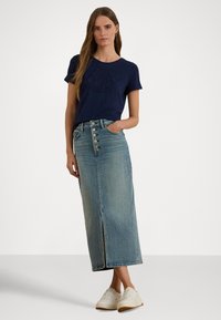 Γυναίκα με μπλε σκούρο κοντομάνικο πουκάμισο και τζιν μίντι φούστα ψηλόμεση με κουμπιά μπροστά, φορώντας λευκά αθλητικά παπούτσια, στέκεται μπροστά σε λευκό φόντο.