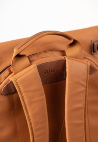 Zaino in pelle marrone con spallacci imbottiti, una toppa con logo e superficie liscia. Presenta cuciture resistenti e una cerniera.