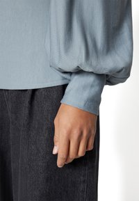 Blusa azzurro chiaro a maniche lunghe con tessuto testurizzato. Presenta una manica ampia e arricciata e un polsino liscio. Abbinata a pantaloni scuri testurizzati.