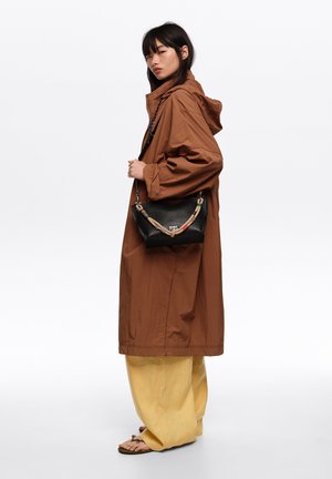 Mujer con un abrigo largo marrón con capucha, pantalones anchos amarillos, sandalias y sujetando un bolso negro texturizado con una correa trenzada de colores.