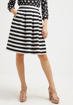 Vrouw draagt een zwart-witte horizontaal gestreepte rok, zwarte blouse met witte hartjes en beige sandalen met hoge hakken, staat met één hand in de zak.