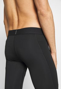 Närbild av en skjortlös person som bär svarta Nike Pro leggings, visar nedre delen av ryggen och övre låren mot en enfärgad bakgrund.