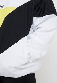Kurtka przeciwdeszczowa z panelami w kolorach czarnym, białym i neonowożółtym; wykonana z gładkiego materiału, z elastycznymi mankietami i logo na piersi.