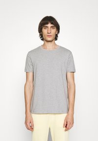 Tricou din bumbac gri, cu guler rotund și mâneci scurte, expus pe un model care poartă pantaloni galbeni deschis. Design simplu, fără modele vizibile.