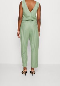 Salopette in lino verde con schiena a V, design senza maniche, gambe affusolate e tasche laterali. Abbinata a sandali neri con tacco.