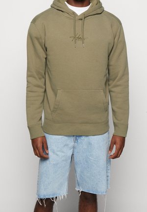 Mies pukeutuneena oliivinvihreään huppariin, jossa on etutasku, ja vaaleansinisiin revittyihin farkkushortseihin, yksinkertaista taustaa vasten.