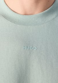 Camiseta de gola redonda azul claro com o lettering em relevo "HUGO" centrado no peito, abaixo do pescoço da pessoa.