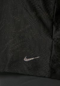 Černý sportovní top s texturovanými vzory a stříbrným reflexním logem Nike na spodní části. Hladký povrch s mírným elastickým efektem.