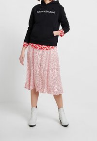 Felpa nera con il testo "CALVIN KLEIN JEANS", blusa rossa a fiori sotto, gonna bianca a fiori e stivaletti bianchi alla caviglia con un design testurizzato.