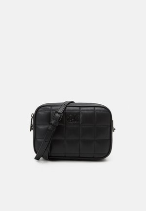 Sac bandoulière matelassé noir avec une bandoulière réglable et un logo "CK" à l'avant sur un fond uni clair.
