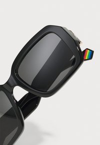 Occhiali da sole neri oversize con lenti scure, caratterizzati da una montatura spessa e lucida. Accentuati da un piccolo dettaglio arcobaleno su una delle tempie.