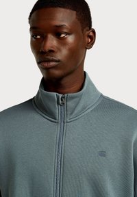 Szara kurtka zapinana na zamek błyskawiczny z wysokim kołnierzem, wykonana z miękkiej tkaniny. Posiada subtelne logo na piersi oraz gładki detal zamka.