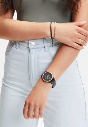 Montre noire avec un cadran noir rond, des accents en or rose et un bracelet en métal noir. Portée avec deux bracelets sur un fond en denim clair.