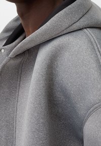 Grijze capuchontrui met een zachte textuur, voorzien van een zwarte voering, metalen drukknoopsluiting en opvallende stikdetails.