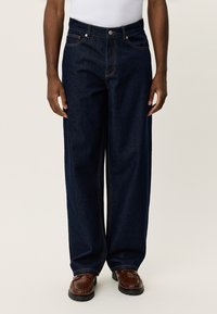 Σκούρο μπλε τζιν παντελόνι με φαρδιά πατζάκια, πορτοκαλί ραφές και κλείσιμο με κουμπιά μπροστά. Φορεμένο με καφέ παπούτσια με υφή.