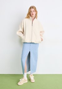 Kreemivärvi fliisist zip-up jakk, helesinine denimpoolik ees oleva lõhega, beežid sneaksid dekoratiivsete paeltega, mudel seisab rohelisel põrandal.
