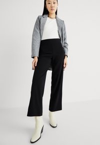 Blazer cinza ajustado, top branco justo, calças largas pretas de cintura alta e botins de salto em tom off-white contra um fundo simples.