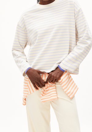 Personne portant une chemise à manches longues rayée jaune pâle et blanche, avec une chemise à rayures pêche et blanches attachée autour de la taille, associée à un pantalon crème.