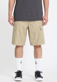 Pantalones cortos de carga beige con bolsillos laterales, fabricados en una mezcla de algodón, combinados con zapatillas negras y calcetines blancos de caña baja.