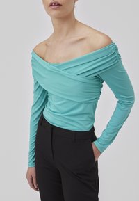 Turkis off-shoulder top med samlet detaljer, lange ærmer og blød tekstur, parret med sorte tætsiddende bukser.