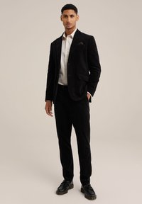 Zwarte corduroy pak met een op maat gemaakte blazer en bijpassende broek. Witte shirt eronder, gecombineerd met zwarte veterschoenen en een pochet.