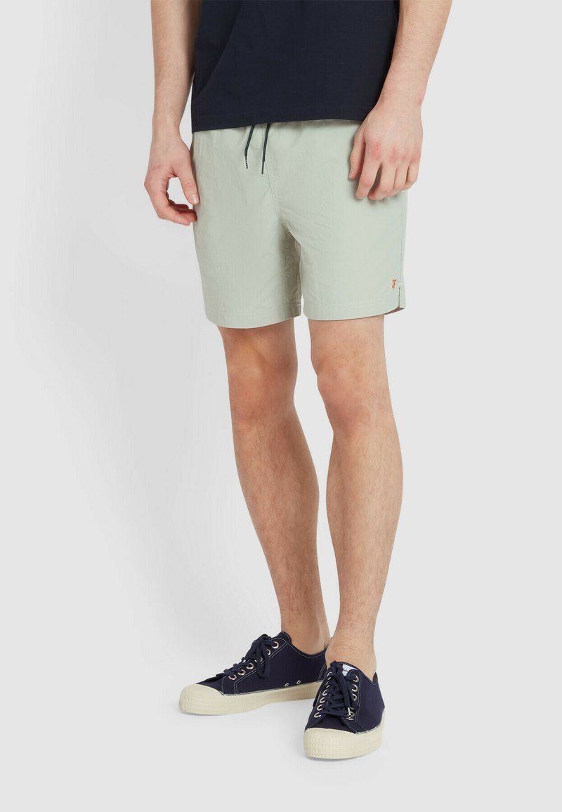 Costumi da bagno verde chiaro con elastico in vita e coulisse, con un accento sul bordo laterale. Abbinati a scarpe da ginnastica blu scuro e una maglietta scura.
