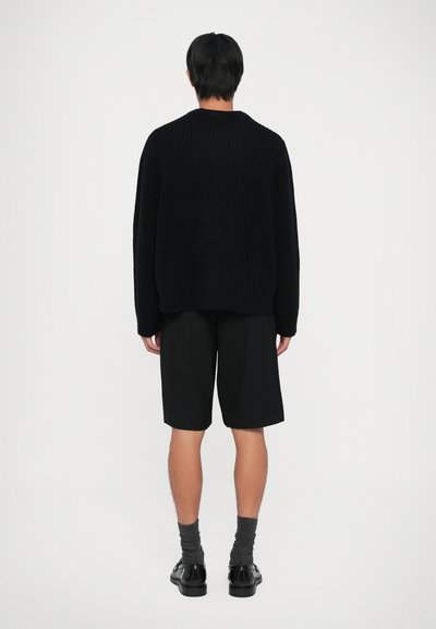 Persona in piedi di spalle che indossa un maglione nero di maglia, pantaloni corti neri fino al ginocchio, calze grigie scure e scarpe eleganti nere su sfondo bianco.