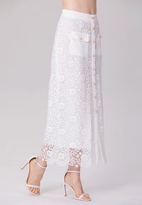 Jupe midi en dentelle blanche avec des motifs floraux élaborés, dotée de boutons sur le devant et de deux poches plaquées. Associée à des talons blancs à lanières.