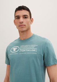 Teal bomulds t-shirt med korte ærmer, rund hals og hvide tekstgrafikker inklusive mærket "TOM TAILOR." Blød tekstur, afslappet design.