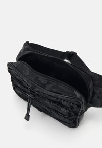 Svart nylonveske med et teksturert, preget mønster, flere glidelåslommer og en justerbar stropp for sikker bæring.