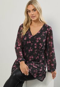 Blusa nera floreale con maniche lunghe, motivo a fiori rosa, scollatura a V, dettaglio in vita arricciato e tessuto trasparente. Indossata con leggins neri.