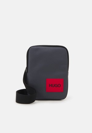 Sac d'épaule en tissu gris à texture lisse, avec un patch logo rouge affichant "HUGO" et une sangle noire réglable. Forme rectangulaire, fermeture éclair.