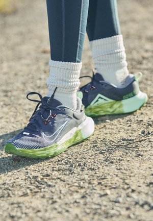 Osoba nosząca czarne i zielone buty do biegania Nike z białymi skarpetkami i niebieskimi legginsami stojąca na żwirowej nawierzchni na zewnątrz.