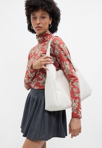 Bolsa tote branca com uma forma estruturada e textura suave, segurada com uma mão. Estampa floral proeminente numa blusa de gola alta vermelha e uma saia plissada cinza.
