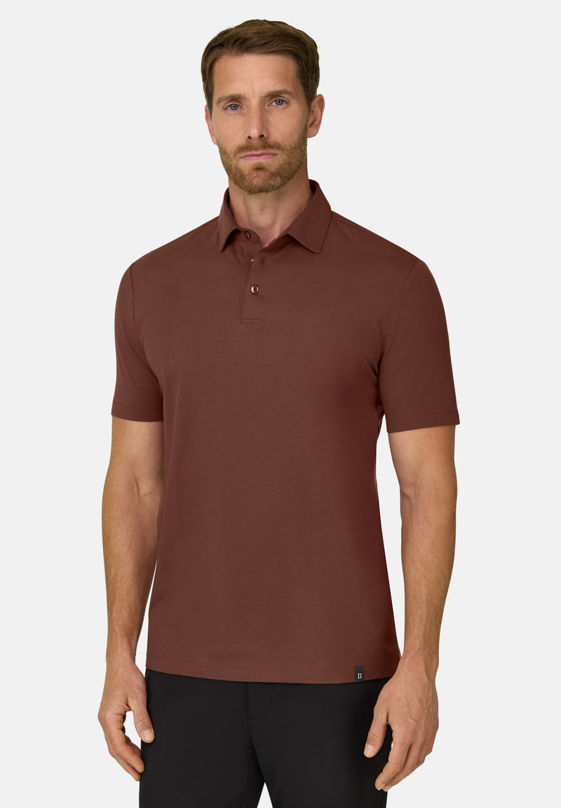 Homme aux cheveux courts et bruns avec une barbe, portant un polo marron à manches courtes et un pantalon noir, debout devant un fond uni.