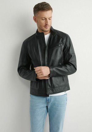 Mężczyzna poprawiający mankiet czarnej skórzanej kurtki, ubrany w jasnoniebieskie dżinsy i białą koszulę, stoi na tle gładkiej jasnej ściany.