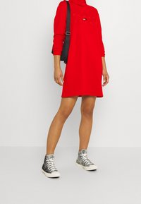 Vestido sudadera rojo con cuello alto y mangas largas, que presenta el texto dorado "TOMMY JEANS". Combinado con zapatillas de caña alta de patrón en blanco y negro.