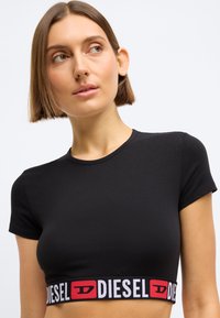 Svart kortermet crop top laget av glatt stoff, med en kontrasterende elastisk midjebånd med den dristige "DIESEL"-logoen i hvitt og rødt.