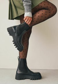 Fekete bőrből készült bokacsizma elasztikus oldalsó panelekkel és vaskos, éles talppal. Fekete szívmintás harisnyával és zöld szoknyával viselve.