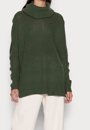 Maglione in maglia verde scuro di grandi dimensioni con colletto a cowl. Presenta un tessuto testurizzato e polsini a coste, abbinato a pantaloni di colore chiaro.