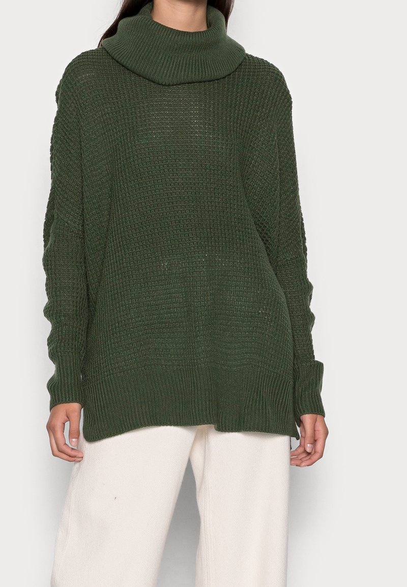 Pull en tricot de grande taille, de couleur vert foncé, avec un col cheminée. Présente un tissu texturé et des poignets côtelés, assorti à un pantalon de couleur claire.