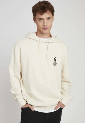 Sweat à capuche crème en tissu doux avec une poche avant ; présente un design graphique noir sur la poitrine gauche. Superposé sur une chemise blanche.