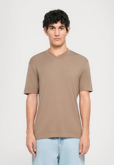 Junger Mann mit lockigem dunklem Haar, der ein braunes kurzärmliges V-Ausschnitt-Shirt und hellblaue Jeans trägt, im Vordergrund vor einem schlichten Hintergrund stehend.