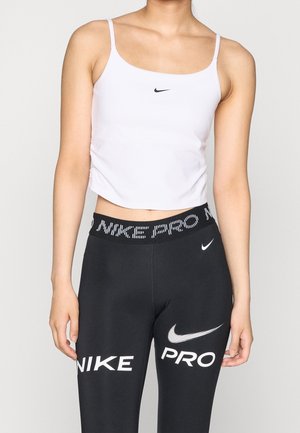 Kobieta ubrana w biały top Nike i czarne legginsy Nike Pro z widocznymi logo marki na ubraniu na jednolitym tle.