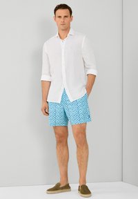 Bijela lanena košulja s dugmadi i podvrnutim rukavima, plavi kupaćih šortsevi s uzorkom i maslinasto zelene slip-on cipele, stoji naslonjen na neutralnu pozadinu.